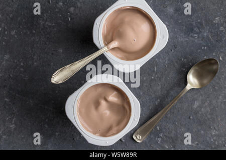 Due aromatizzato al cioccolato yogurt in bicchieri di plastica con cucchiai su grigio texture di sfondo - top view foto di tazze di yogurt Foto Stock