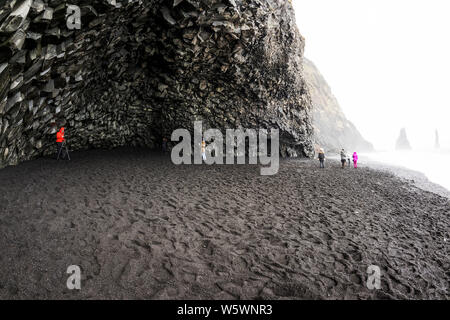 I turisti ad esplorare rare grotta rocciosa formazioni in la spiaggia di sabbia nera, Islanda Foto Stock