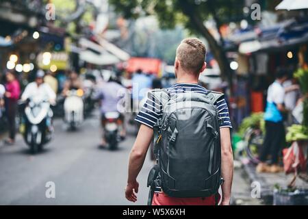 Traveler camminando sulla strada trafficata. Vista posteriore del giovane con uno zaino nel quartiere vecchio di Hanoi, Vietnam. Foto Stock