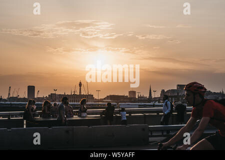 London, Regno Unito - Luglio 23, 2019: i pedoni e i ciclisti sul Ponte di Londra durante il tramonto, motion blur, treni e dello skyline della città sullo sfondo. Londra è Foto Stock