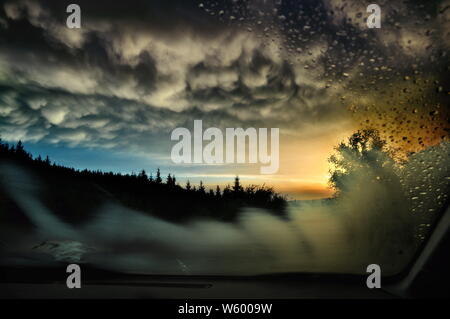 Un tramonto in una zona di montagna, visibile attraverso il parabrezza di un auto, su cui le gocce di pioggia e la cottura a vapore sono visibili. Foto Stock