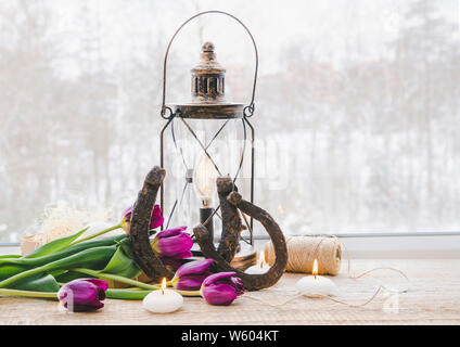 Vecchi ferri di cavallo arrugginito, marrone vintage lanterna con moderni decorativi dimmerabile lampadina accesa e candele bianche accese e tulipani sulla finestra in legno Foto Stock