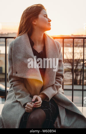 Caucasian i capelli castani ragazza in un cappotto beige si siede nel vento e guarda al lato. La luce del tramonto dietro Foto Stock