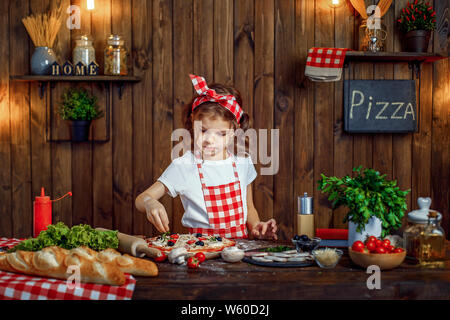 Ragazza sorridente indossando T-shirt bianca con grembiule a scacchi archetto e spruzza la pizza con verdi sulla tabella riempita con gli ingredienti per la pizza in elegante cucina in legno Foto Stock