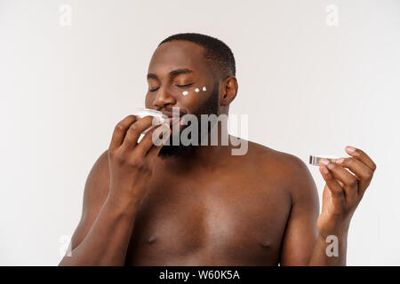 Sorridente uomo afro applicare la crema sul suo volto. Uomo di cura della pelle del concetto. Foto Stock