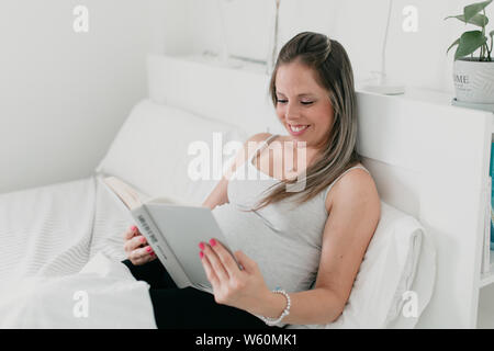 Incinta donna bionda che giace nel letto la lettura di un libro Foto Stock