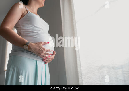 Donna incinta guarda fuori dalla finestra mentre si tocca la sua pancia Foto Stock