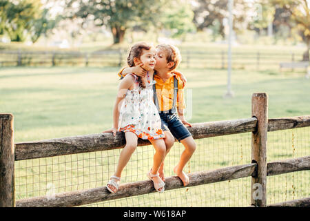 Due simpatici divertenti bambini caucasici ragazzo ragazza seduta sul recinto nel parco al di fuori. Amici fratelli kids abbracciando il bacio al tramonto d'estate. Amore romantico e Foto Stock