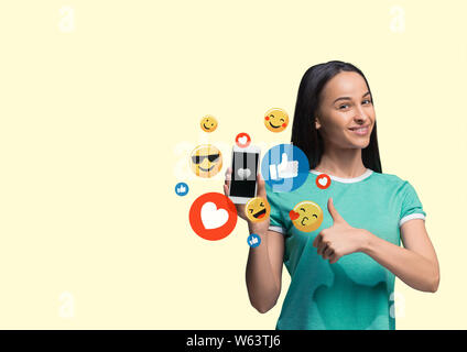 I social media le interazioni sul telefono cellulare. Internet marketing digitale, Chating, commentando e gradimento. Sorrisi e le icone sopra lo schermo dello smartphone, l'azienda dalla giovane donna di colore giallo su sfondo per studio. Foto Stock