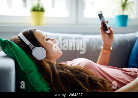 Vista laterale di una giovane donna rilassante sul divano di casa, l'ascolto di musica con cuffie e telefono azienda Foto Stock