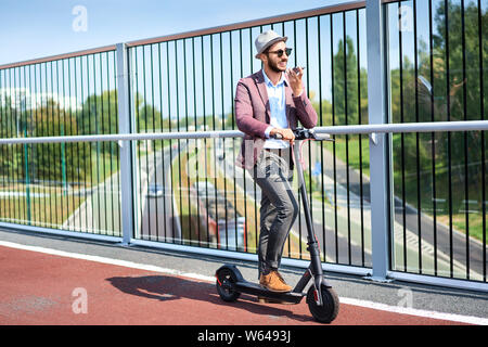 Giovane uomo in piedi su scooter motorizzato nella città e parlando al telefono Foto Stock