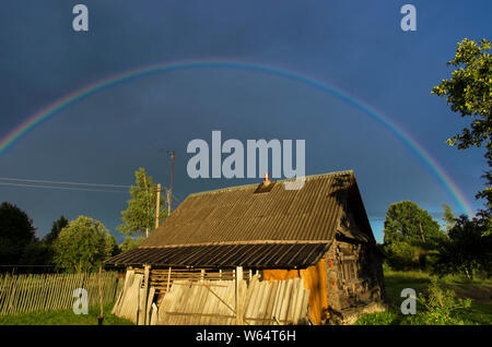 Arcobaleno nel cielo drammatico su un vecchio imbarcati log casa di campagna Foto Stock