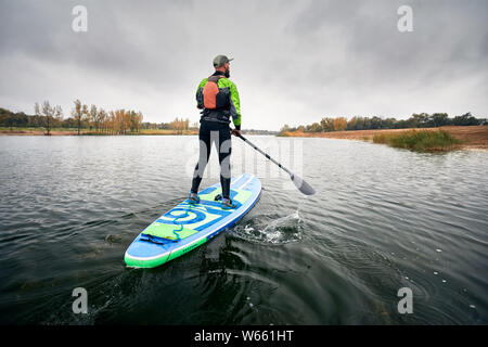 Atleta in neoprene paddleboard ad esplorare il lago a freddo contro il cielo nuvoloso Foto Stock