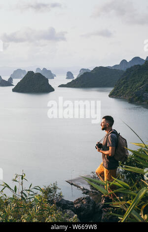 Uomo caucasico con una telecamera in piedi su una scogliera che si affaccia sulla baia di Halong durante il tramonto, Vietnam. Orientamento verticale Foto Stock