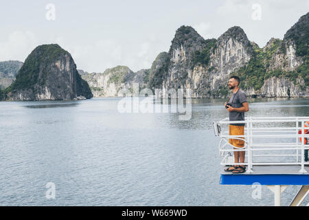 Giovane uomo caucasico con una telecamera in piedi su un ponte di traghetto affacciato sulla baia di Halong, Vietnam. Foto Stock