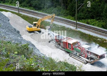 Versam, GR / Svizzera - 30. Luglio 2019: escavatore e rock macinatore al lavoro per produrre la massa fini ciottoli di grandi rocce accanto ai binari della ferrovia Foto Stock