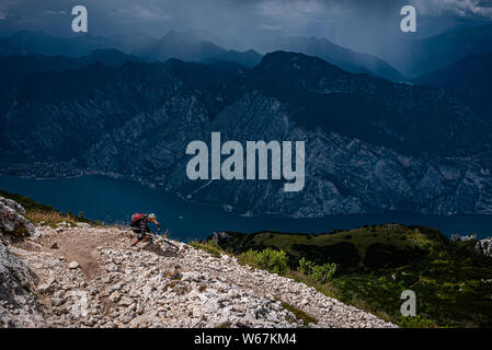 Il LAGO DI GARDA, Italia. Un mountain biker in sella ad un angolo su un pericoloso sentiero roccioso alto sopra il Lago di Garda con scuri sensazionali montagne e nuvole di tempesta in Foto Stock