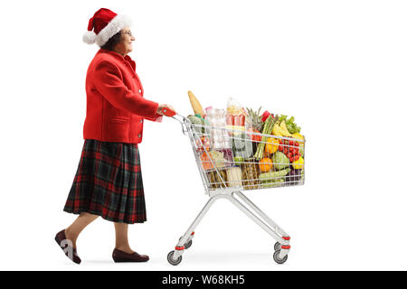 Lunghezza piena ripresa del profilo di una donna anziana con un Natale spingendo un carrello della spesa con prodotti alimentari isolati su sfondo bianco Foto Stock