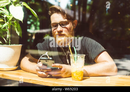 Attraente redhead con barba ascolta la musica su un telefono cellulare mentre seduti a un tavolo del bar. Sorridente maschio utilizza uno smartphone Foto Stock