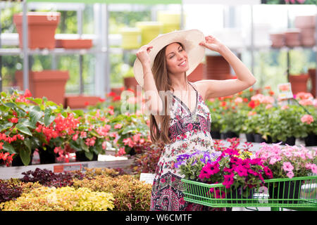 Felice elegante ragazza shopping per vasi di piante fiorite in un vivaio in un grazioso abiti estivi e ampia colmato cappello sorridente come lei regola la Foto Stock