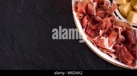 La birra snack e tapas: formaggio, prosciutto sulla piastra bianca su sfondo scuro. Alimenti e bevande Foto Stock