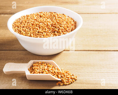 Fenugreek (Trigonella) in una ceramica tazza bianca con scoop su uno sfondo di legno Foto Stock