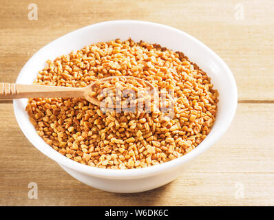 Fenugreek (Trigonella) in una ceramica tazza bianca con cucchiaio su uno sfondo di legno Foto Stock