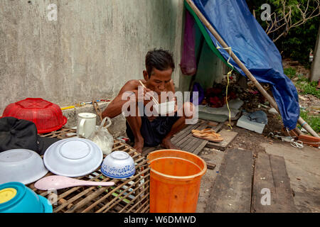 Un anziano senzatetto cambogiano è di mangiare un pasto vicino alla sua casa sgangherato su una strada di città in Kampong Cham, Cambogia. Foto Stock