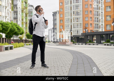 Giovane uomo con zaino sulle spalle di parlare al telefono tenendo black coffee cup sullo sfondo di case multipiano. Studente guardando le case con architettura moderna. Foto Stock