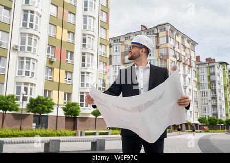 Giovane ingegnere in bianco hardhat in piedi con la costruzione architettonica disegni, guardando case multipiano. Lavoratore che posano su sfondo di appartamenti nuovi edifici. Foto Stock