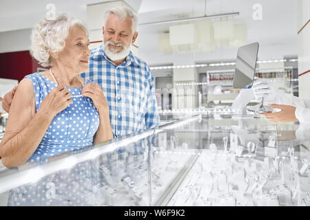 Allegro barbuto senior Uomo in camicia a scacchi abbracciando la sua bella moglie in vestito blu che cercando su Collana di perle in gioielleria. Capelli grigi marito piacevole facendo presente alla donna amata Foto Stock