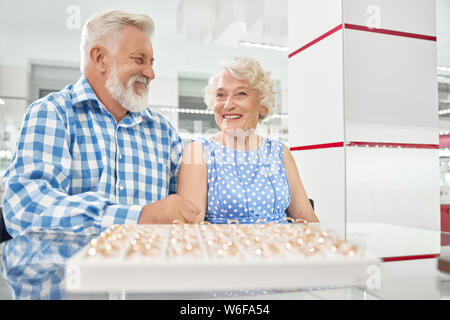 Sorridente piuttosto alti lady in polka dot dress è venuto al negozio di lusso con la sua amata dai capelli grigi marito per selezionare gioielli esclusivi. Felice Coppia matura godendo di tempo di shopping insieme Foto Stock