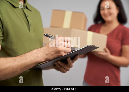 Close up di consegna uomo in verde t shirt tenendo la cartella con i documenti e scrivere con la penna. Allegro, donna positiva ricezione ordine in due scatole di cartone. Foto Stock