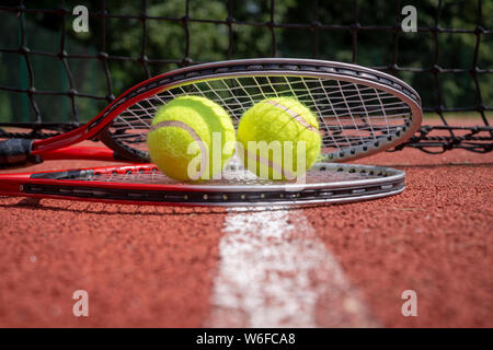 Campi da tennis in scena con il nero net, palle da tennis in appoggio sulla sommità di una racchetta da tennis su red hard court superficie, a basso angolo di visione e la messa a fuoco selettiva Foto Stock