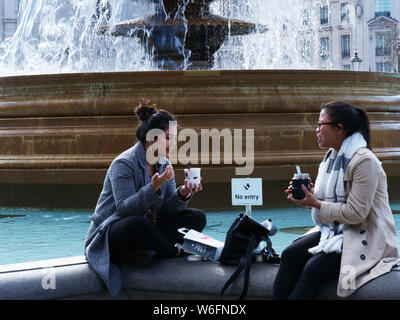 Due donne aventi il pranzo e parlare mentre seduto sul bordo della vasca che circonda la fontana ovest in Trafalgar Square, Londra, nel mese di marzo Foto Stock