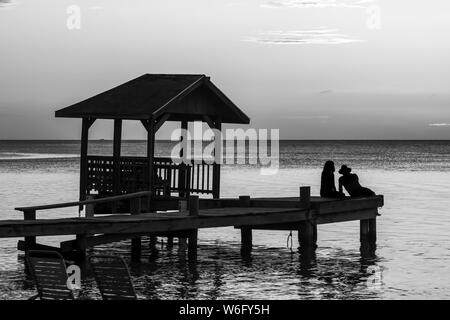 Una coppia si siede alla fine di un molo che si affaccia sul tranquillo oceano al tramonto; Roatan, Bay Islands Department, Honduras Foto Stock