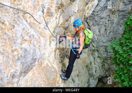 Turista femminile con ingranaggio di arrampicata e zaino in appoggio su una via ferrata, appeso ad un gancio di bloccaggio attaccato al suo set da via ferrata da t Foto Stock