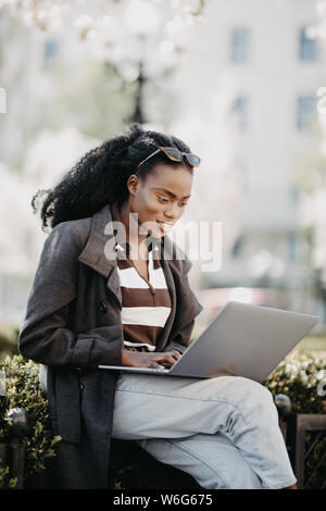 Bella black business donna seduta sul banco nella città finanziaria, utilizzando un computer portatile, premuroso, soleggiata all'esterno. Foto Stock