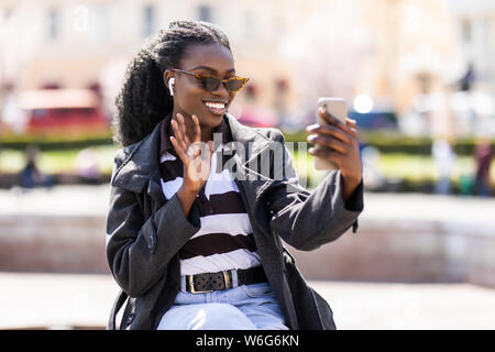 Ritratto closeup di amichevole americano africano Donna con cuffie rendendo la chiamata video sullo smartphone mentre è seduto sul banco vicino al lungofiume Foto Stock