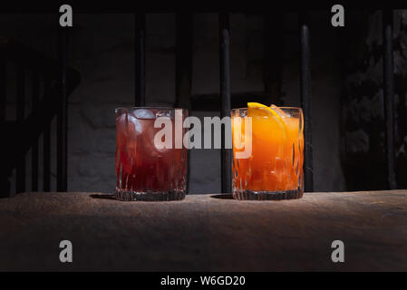 Due cocktail colorati nella barra di colore scuro. L'arancione e il rosso alcol bevande con ghiaccio su un tavolo di legno. Maschio e femmina di bere in eleganti occhiali. Foto Stock