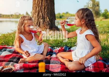 Sorelle di bere succo di frutta mentre avente picnic sulle rive del fiume estate al tramonto. I bambini a mangiare la torta e rilassante con la famiglia all'aperto Foto Stock