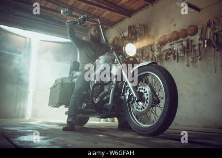 Bello brutale con barba seduto su una moto nel suo garage. Foto Stock