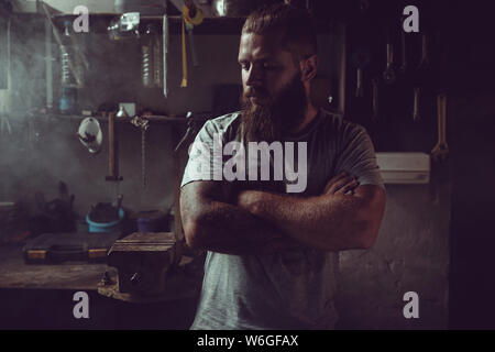 Bello brutale con barba in piedi nel suo garage contro lo sfondo di strumenti di riparazione. Foto Stock