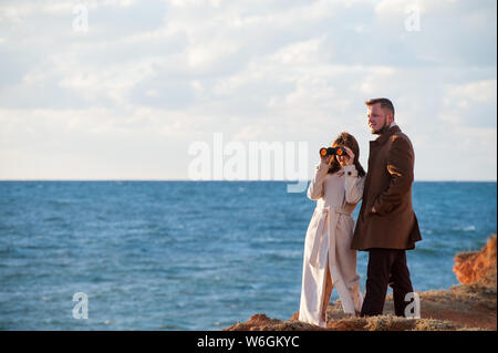 Felice bel giovane coppia alla moda uomo e donna in autunno ricoprire guardando attraverso il binocolo vicino al mare con copyspace Foto Stock