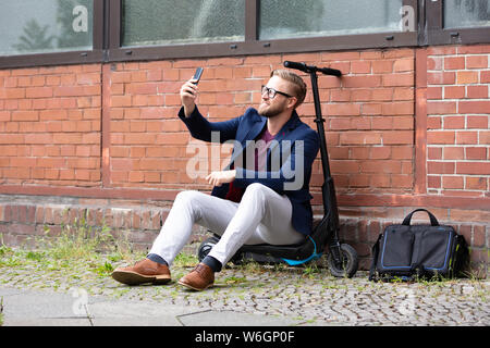 Imprenditore sorridente seduto su scooter elettrico vicino a un muro di mattoni tenendo Selfie con telefono mobile Foto Stock