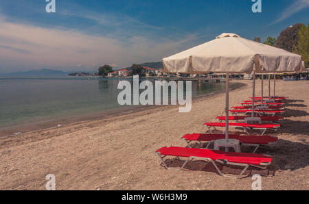 Volos, Magnesia, Grecia - 26 Aprile 2015: fila di lettini rosso su una spiaggia tranquilla con alcune case nella parte inferiore dell'immagine Foto Stock