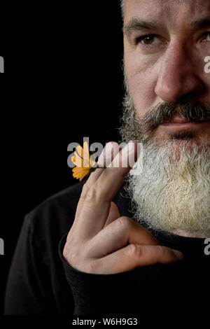 Closeup ritratto di una brutale dai capelli grigi con barba con un fiore giallo nelle sue mani su uno sfondo nero, il fuoco selettivo Foto Stock