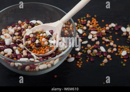Fagioli, lenticchie, Mung bean e piselli in una ciotola di vetro e il cucchiaio di legno su uno sfondo nero. Concetto - Dieta cheto. Foto Stock