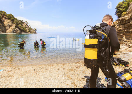 Vista posteriore di scuba diver uomo con serbatoio di ossigeno la preparazione per le immersioni . Gruppo di sub al mare in background. Estate attività estreme . Foto Stock