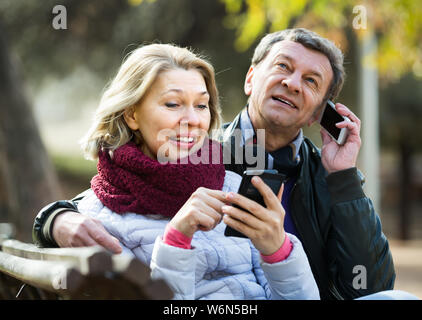 I pensionati gioiosa giovane occupato con telefoni cellulari durante passeggiate al parco. Focus sulla donna Foto Stock